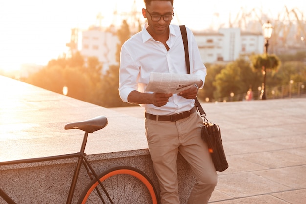 Beau jeune homme africain en début de matinée avec vélo lisant le journal.