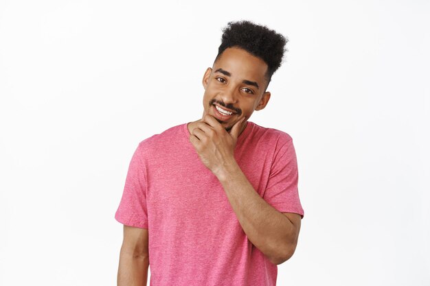 Beau jeune homme afro-américain avec une coupe de cheveux élégante, touchant sa barbe et des dents blanches souriantes, debout dans un t-shirt rose décontracté sur fond blanc