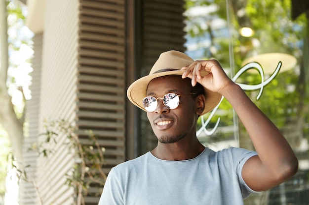 Photo gratuite beau jeune homme afro-américain souriant portant des lunettes rondes en miroir, ajustant son chapeau beige, ayant un regard insouciant et heureux, attendant son déjeuner tout en se détendant à la cafétéria en plein air