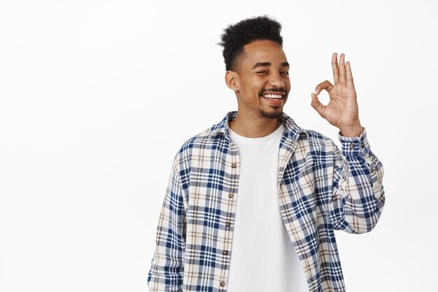 beau jeune homme afro-américain souriant satisfait, montrant OK, OK signe zéro et hoche la tête en signe d'approbation, note une bonne chose, loue un excellent travail, recommande le magasin, debout sur blanc