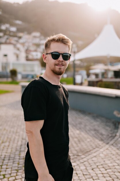 Beau jeune homme aux cheveux blonds portant un t-shirt noir et des lunettes de soleil marche sur la ville carrée au coucher du soleil L'homme voyage sur le rivage de l'océan