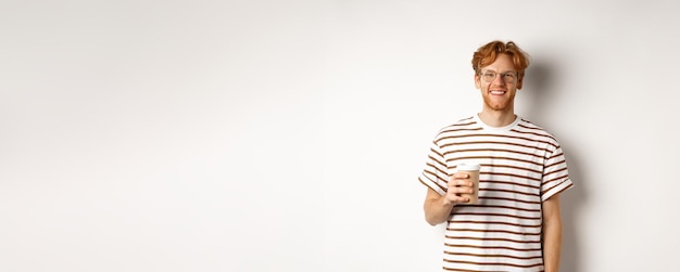 Photo gratuite beau jeune homme à la barbe et aux cheveux rouges en désordre portant des lunettes avec un t-shirt rayé buvant du café