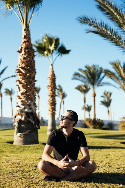 Beau jeune homme barbu à lunettes de soleil assis sur l'herbe sous les palmiers en vacances d'été luxary resort