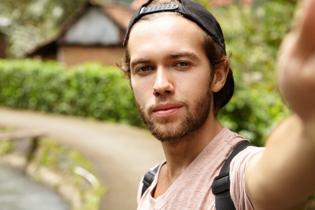 Beau jeune homme barbu portant une casquette de baseball en arrière prenant selfie, regardant avec sourire, posant sur route de campagne contre la nature verte