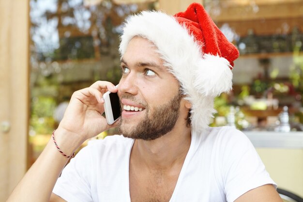 Beau jeune homme barbu portant un chapeau de père Noël à l'aide de téléphone mobile générique