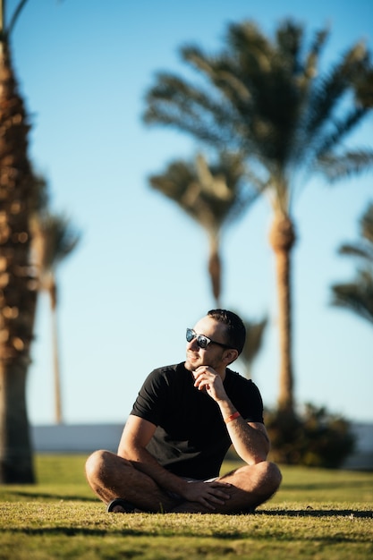 Beau jeune homme barbu regardant de côté dans des lunettes de soleil assis sur l'herbe sous les palmiers en vacances d'été.