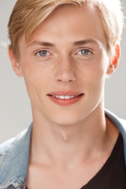 Beau jeune homme blond souriant portant un t-shirt noir décontracté et une veste en jean sur un mur gris