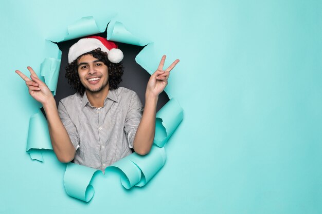 Beau jeune homme bouclé porter en bonnet de noel avec geste de paix du trou sur du papier vert