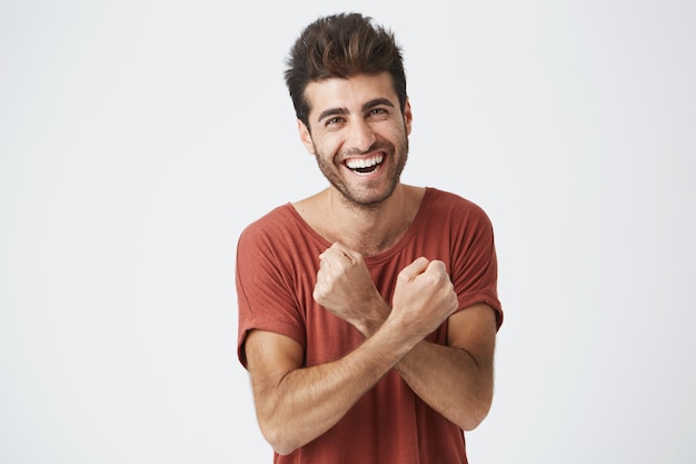 Beau jeune homme bronzé se sentant excité, faisant des gestes activement, gardant les poings serrés et croisés, riant joyeusement, heureux avec bonne chance. Une étudiante excitée par la réussite des examens