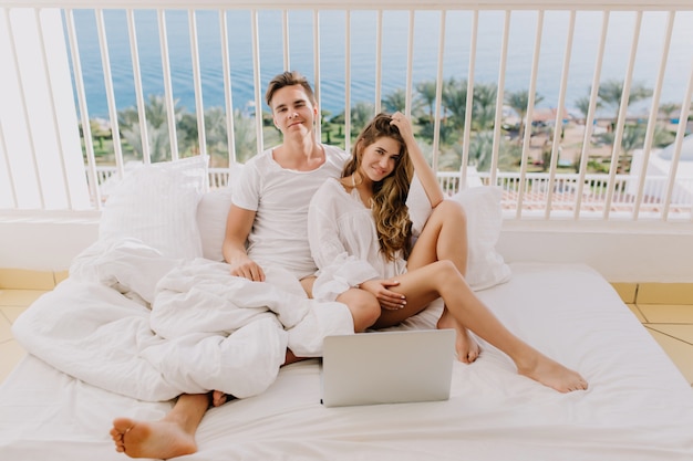 Beau jeune homme en chemise blanche souriant tandis que sa jolie petite amie rêveuse touchant ses longs cheveux. Beau couple, passer du temps avec un ordinateur portable posant