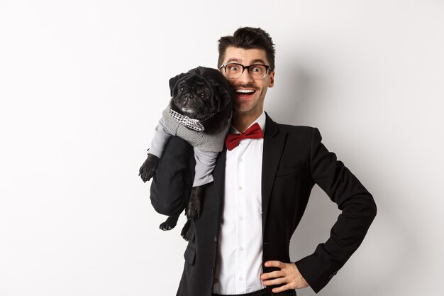 Beau jeune homme en costume et lunettes tenant un mignon chien carlin noir sur l'épaule, souriant heureux devant la caméra, portant des tenues de fête, fond blanc.