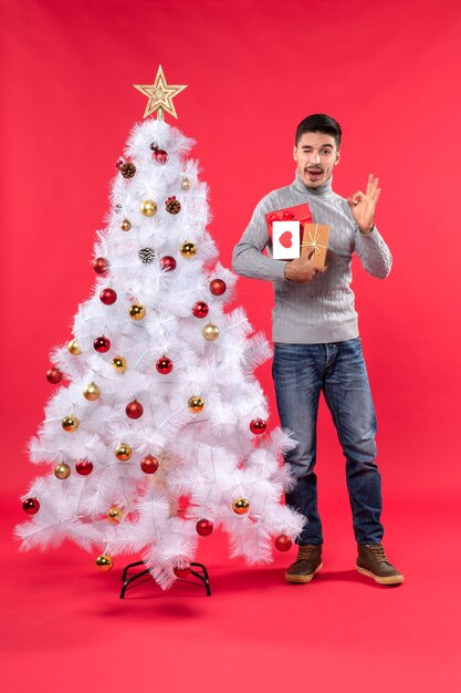 Beau jeune homme debout près de l'arbre du Nouvel An blanc décoré et tenant ses cadeaux