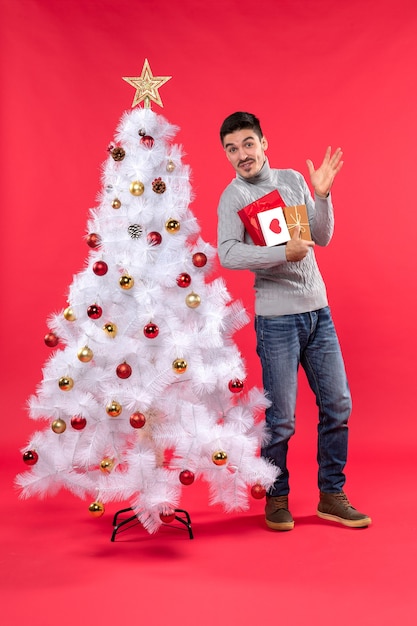 Beau jeune homme drôle debout près de l'arbre du Nouvel An blanc décoré et tenant ses cadeaux