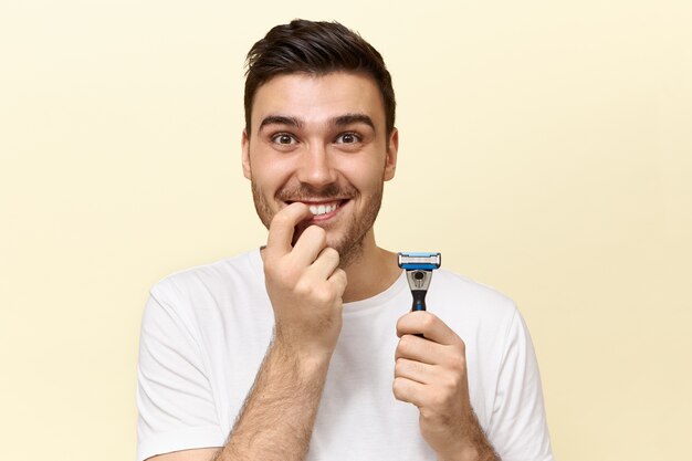 Beau jeune homme drôle émotionnel avec poils posant isolé avec bâton de rasage et se ronger les ongles