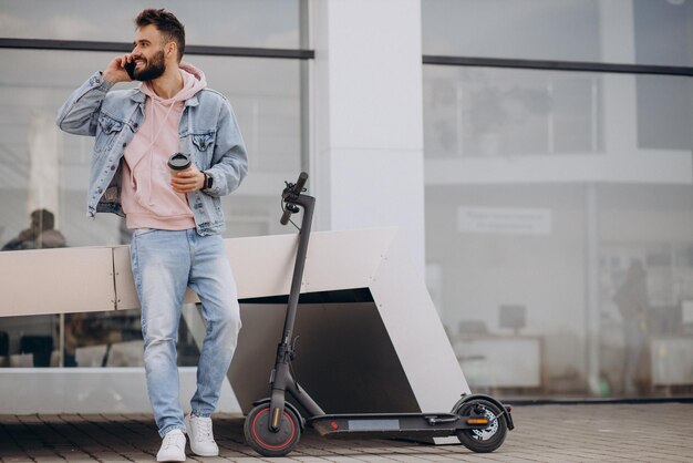 Beau jeune homme faisant du scooter électrique en utilisant le téléphone et en buvant du café