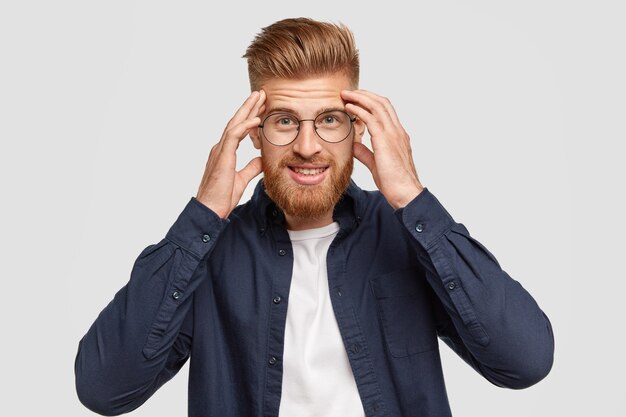 Beau jeune homme gingembre garde les mains sur les tempes, essaie de se concentrer sur quelque chose, se tient contre un mur blanc, a un sourire doux, satisfait des résultats du travail, trouve une solution à l'esprit