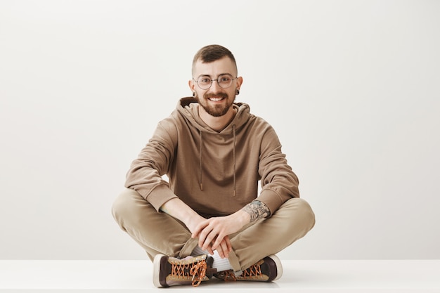 Beau jeune homme à lunettes assis sur le sol et souriant amical