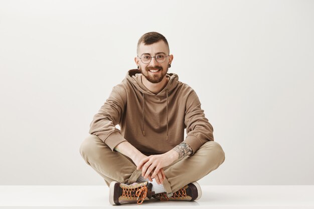 Beau jeune homme à lunettes assis sur le sol et souriant amical