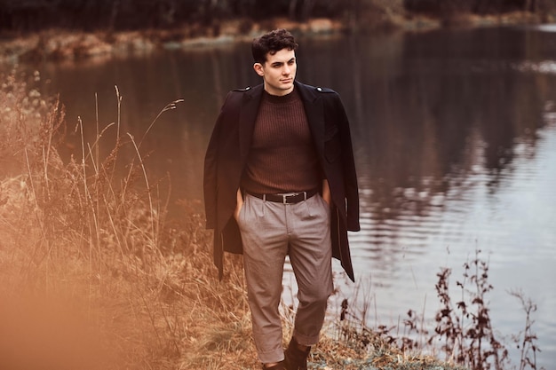 Photo gratuite un beau jeune homme avec un manteau couvrant ses épaules tout en se tenant près d'un lac dans la forêt d'automne.