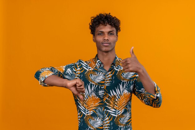 Un beau jeune homme à la peau foncée aux cheveux bouclés en chemise imprimée de feuilles montrant les pouces de haut en bas