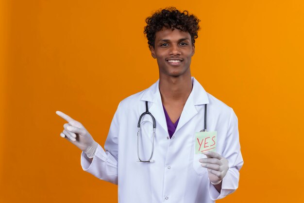 Un beau jeune homme à la peau foncée aux cheveux bouclés portant un manteau blanc avec stéthoscope montrant une carte papier avec le mot oui