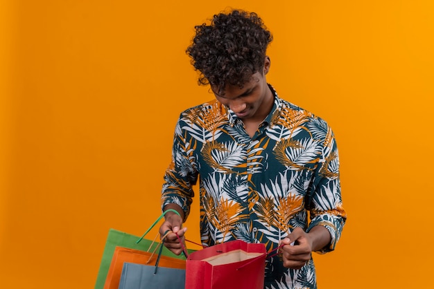 Un beau jeune homme à la peau sombre aux cheveux bouclés en chemise imprimée de feuilles à la recherche de sacs