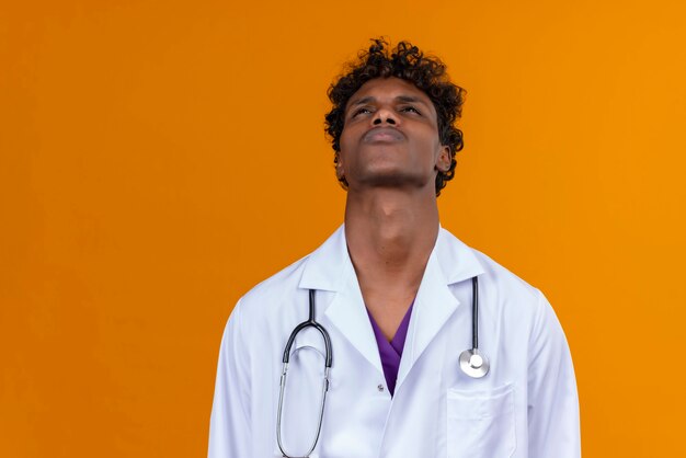 Un beau jeune homme à la peau sombre aux cheveux bouclés portant un manteau blanc avec stéthoscope à tristement