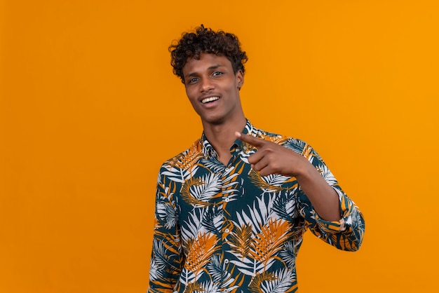 Photo gratuite un beau jeune homme à la peau sombre avec des cheveux bouclés en chemise imprimée de feuilles souriant et pointant avec l'index