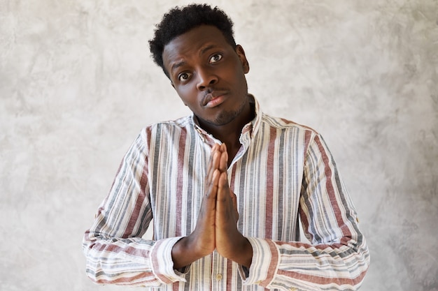 Beau jeune homme à la peau sombre mignon gardant les mains pressées ensemble, ayant une expression faciale lugubre, demandant pardon, être désolé, se sentir coupable, ne voulait pas vous blesser