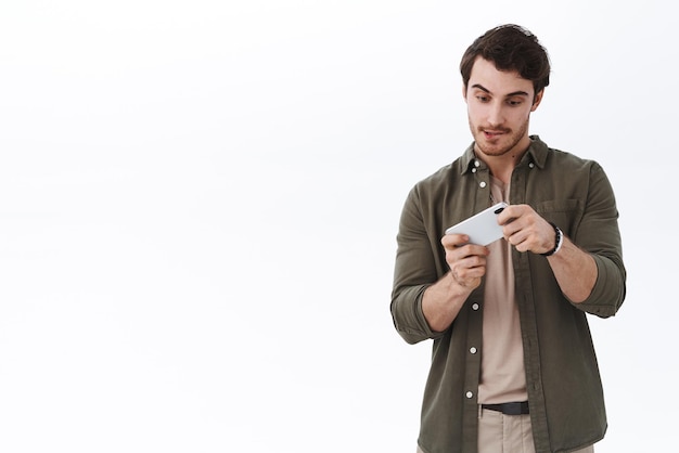 Beau jeune homme de race blanche de 25 ans avec la langue de bâton de moustache comme étant concentré sur la réussite de la mission tout en jouant au jeu de téléphone mobile de niveau difficile tourner le téléphone sur le côté debout fond blanc