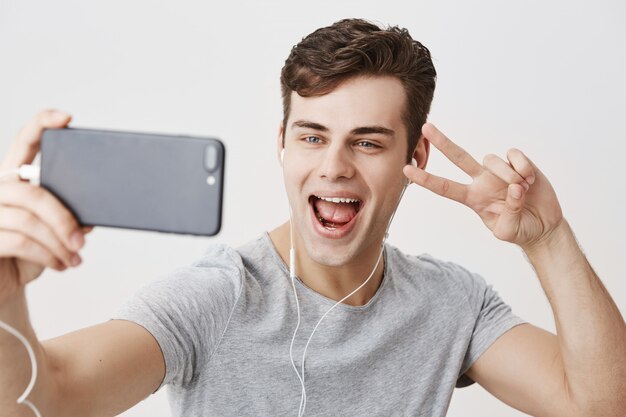 Beau jeune homme de race blanche portant des écouteurs blancs, tenant un téléphone mobile, faisant un appel vidéo, posant pour selfie, souriant largement, montrant le signe V. Communication et technologie modernes.