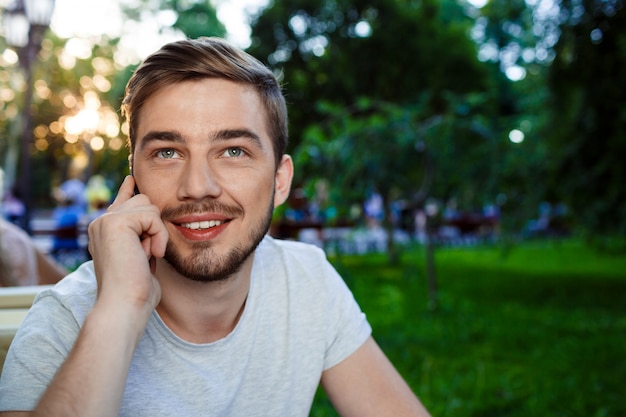 Beau jeune homme souriant assis à la table dans un café en plein air talkin sur téléphone en levant.