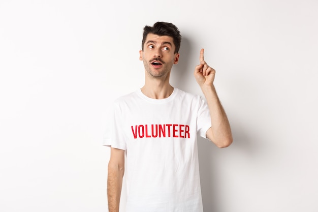 Beau jeune homme en t-shirt bénévole ayant une idée, levant le doigt et disant suggestion, fond blanc.