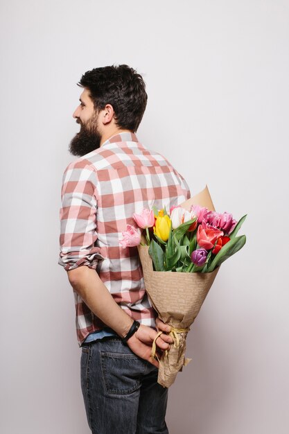 Beau jeune homme tenant un bouquet de fleurs derrière son dos et debout contre un mur blanc