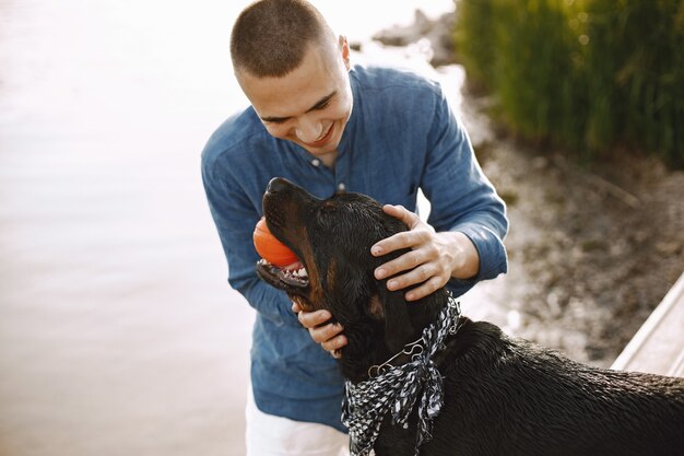 Beau jeune homme en tenue décontractée jouant avec un chien mignon tout en se tenant près du lac. Garçon portant une chemise bleue et un short en jean blanc