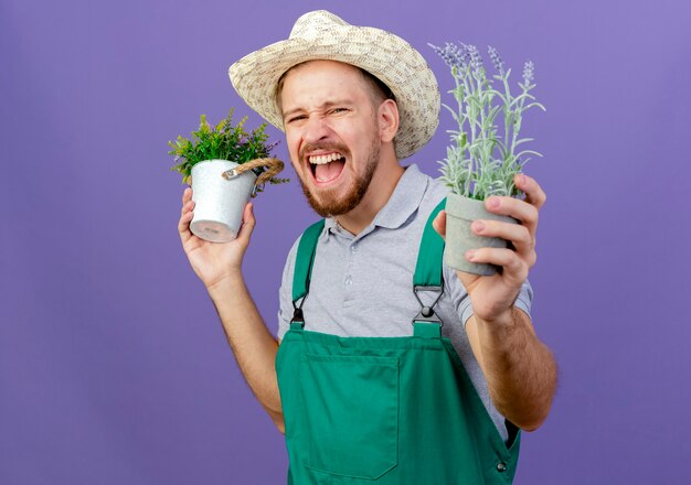 Beau jeune jardinier slave en uniforme et chapeau tenant des pots de fleurs et hurlant isolé sur mur violet avec espace copie