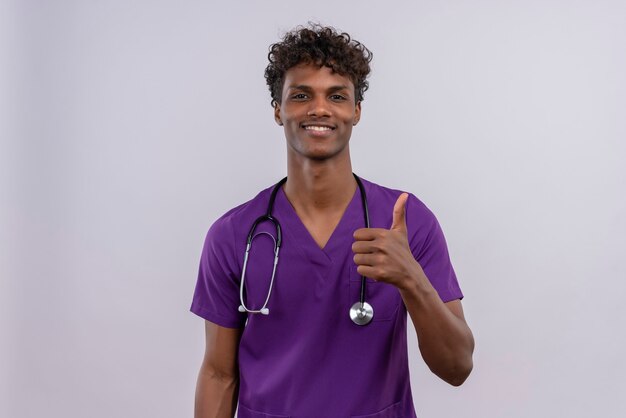 Un beau jeune médecin à la peau sombre avec des cheveux bouclés portant l'uniforme violet avec stéthoscope heureux tout en montrant les pouces vers le haut