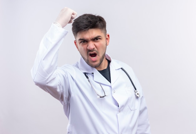 Beau jeune médecin portant une robe médicale blanche, des gants médicaux blancs et un stéthoscope menaçant de poing debout sur un mur blanc