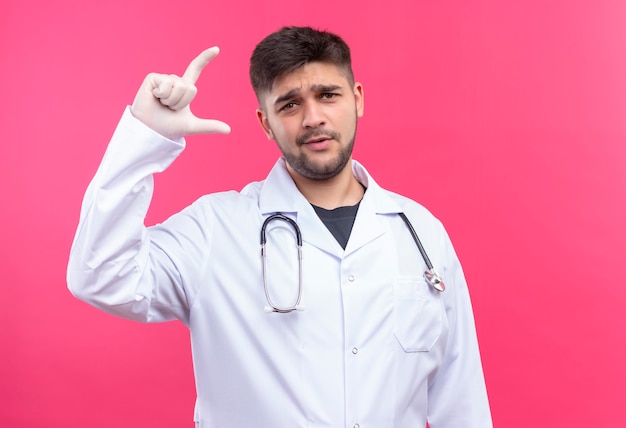 Beau jeune médecin portant une robe médicale blanche gants médicaux blancs et stéthoscope perplexe de mesure de la taille avec les doigts debout sur le mur rose