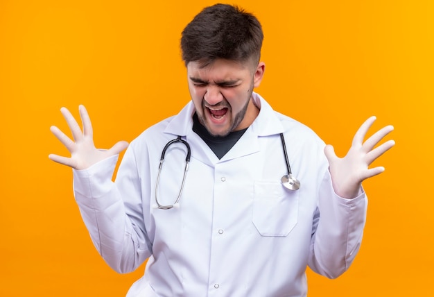 Beau jeune médecin portant une robe médicale blanche, des gants médicaux blancs et un stéthoscope pleurant en colère fermant les yeux debout sur un mur orange