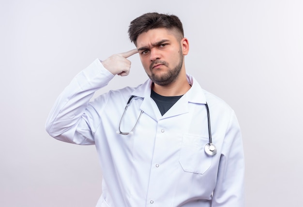 Photo gratuite beau jeune médecin portant une robe médicale blanche, des gants médicaux blancs et un stéthoscope à la recherche de reproche veut que quelqu'un pense debout sur un mur blanc