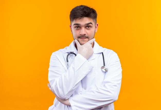 Beau jeune médecin portant une robe médicale blanche, des gants médicaux blancs et un stéthoscope à la smartly debout sur un mur orange