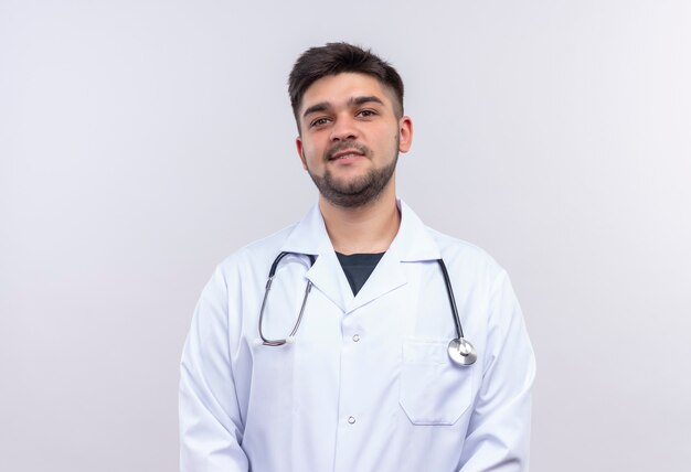 Beau jeune médecin portant une robe médicale blanche, des gants médicaux blancs et un stéthoscope souriant debout sur un mur blanc