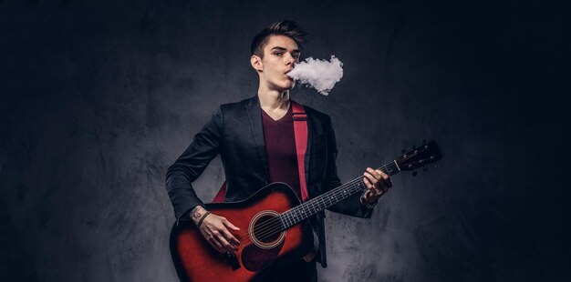 Beau jeune musicien aux cheveux élégants dans des vêtements élégants exhale de la fumée tout en jouant de la guitare acoustique. Isolé sur un fond sombre.