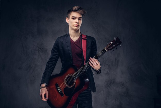Beau jeune musicien aux cheveux élégants dans des vêtements élégants posant avec une guitare dans ses mains sur un fond sombre.