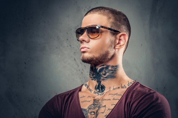 Beau mâle athlétique en lunettes de soleil avec des tatouages sur les bras et le cou posant sur un fond artistique gris dans un studio.