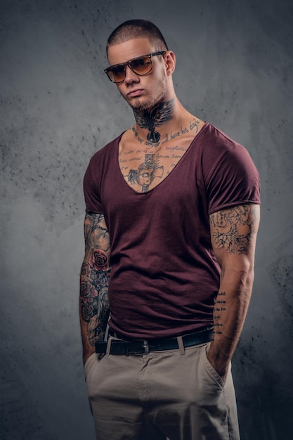 Photo gratuite beau mâle athlétique en lunettes de soleil avec des tatouages sur les bras et le cou posant sur un fond artistique gris dans un studio.