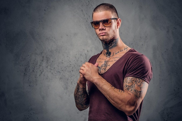 Beau mâle athlétique en lunettes de soleil avec des tatouages sur les bras et le cou posant sur un fond artistique gris dans un studio.