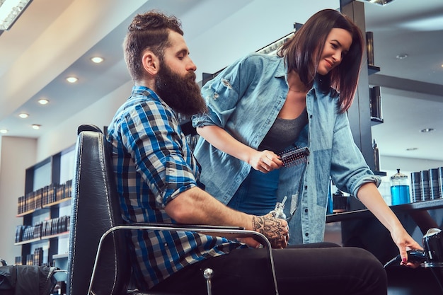 Beau mâle barbu élégant avec un tatouage sur le bras vêtu d'une chemise en flanelle tenant du jus tout en se faisant couper les cheveux.