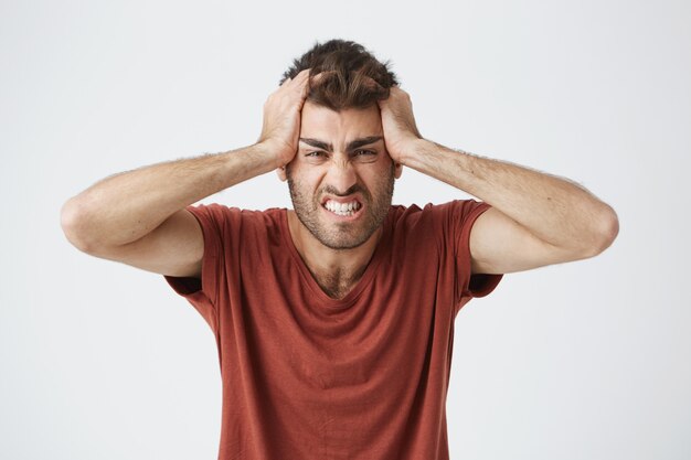 Beau mâle caucasien en colère en t-shirt rouge ayant des expressions folles, serrant la tête avec les mains énervées de gens autour du travail. Émotions négatives.